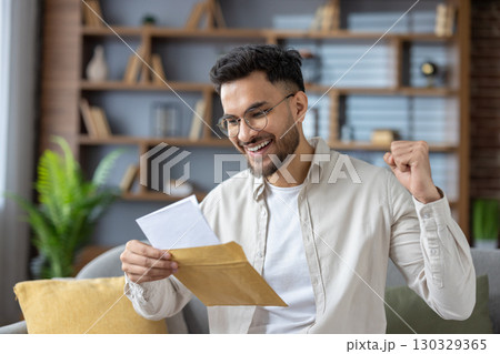 Close-up photo of a young indian man in a shirt and bib sitting on the couch at home and reading a letter he received, happy with the news and message. Close-up photo of a young indian man in a shirt and bib sitting on the couch at home and reading a letter he received, happy with the news and message. 130329365