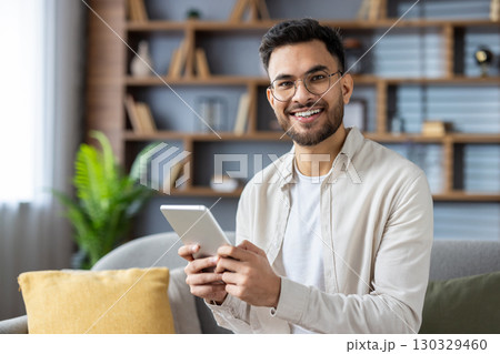 Cheerful man with glasses holding tablet sitting on sofa enjoying digital content. Modern living room setting conveys relaxation and home comfort. Young male engages with technology, showcasing 130329460