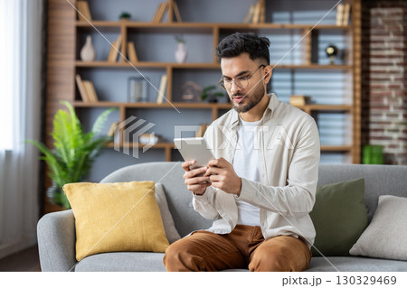 Young man using tablet seated on sofa in cozy living room setting. Concentrated on screen with serious demeanor, immerses in digital reading or online browsing. Young man using tablet seated on sofa in cozy living room setting. Concentrated on screen with serious demeanor, immerses in digital reading or online browsing. 130329469
