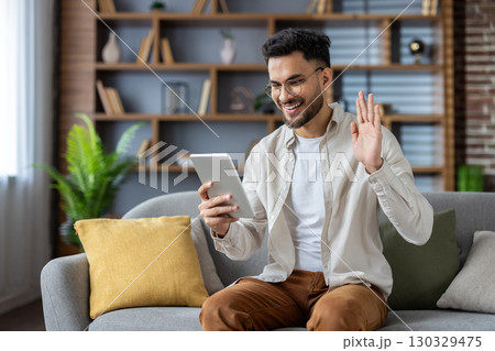 Hispanic man engaged in video call using tablet, sitting on couch, smiling and waving. Captures essence of connection, technology, and communication in modern home setting. 130329475