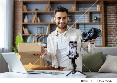 Young man creating unboxing video using smartphone. He holds virtual reality headset and cardboard box, capturing engaging content in home setting. Enthusiastic expression shows excitement about tech Young man creating unboxing video using smartphone. He holds virtual reality headset and cardboard box, capturing engaging content in home setting. Enthusiastic expression shows excitement about tech 130329501