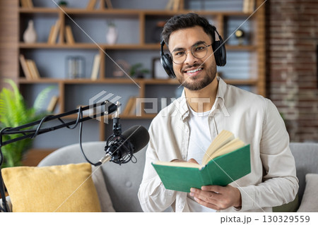 Smiling man wearing headphones recording podcast while reading book. Modern home studio setup with microphone. Engaging in content creation, learning, and sharing information Smiling man wearing headphones recording podcast while reading book. Modern home studio setup with microphone. Engaging in content creation, learning, and sharing information 130329559
