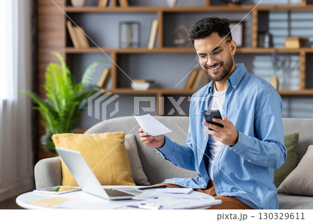Smiling young Indian man sitting on sofa at home, holding documents and bills and looking at mobile phone. 130329611