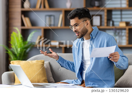 Frustrated and worried young Indian man sitting at home on sofa in front of laptop, holding documents in hand, looking upset at screen. 130329620