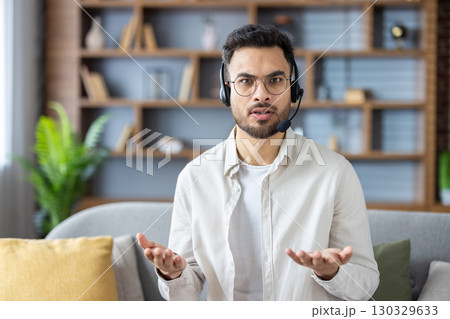 Portrait of a young Indian man wearing glasses and a headset sitting on the couch at home and talking to the camera, while gesturing with his hands. Portrait of a young Indian man wearing glasses and a headset sitting on the couch at home and talking to the camera, while gesturing with his hands. 130329633
