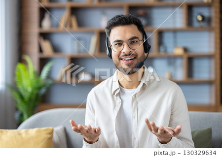 Portrait of a young hispanic man in a headset and glasses sitting on the couch at home and talking to the camera, while gesturing with his hands. Portrait of a young hispanic man in a headset and glasses sitting on the couch at home and talking to the camera, while gesturing with his hands. 130329634