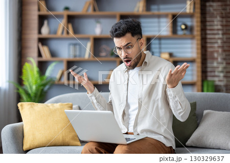 Shocked and worried young muslim man man sitting on sofa at home and dissapointed spreading his hands looking at laptop screen. 130329637