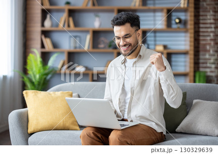 Happy young Indian man sitting on sofa at home, using laptop on lap, looking smiling at screen, showing victory yes with hand. 130329639