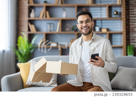 Portrait of a young Muslim man in glasses sitting happy at home on the couch, holding a phone and an open parcel box, looking and smiling at the camera. 130329655