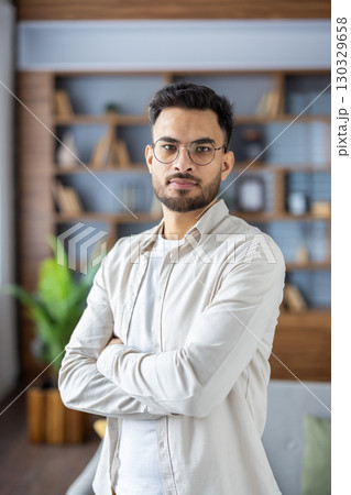 Portrait of a young Indian man wearing glasses standing at home, crossing his arms, standing seriously and confidently looking at the camera. Vertical photo. Portrait of a young Indian man wearing glasses standing at home, crossing his arms, standing seriously and confidently looking at the camera. Vertical photo. 130329658