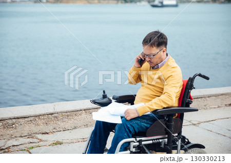 Smiling young disabled man use phone on a wheelchair in a park at sunlight outdoors. Smiling young disabled man use phone on a wheelchair in a park at sunlight outdoors. 130330213
