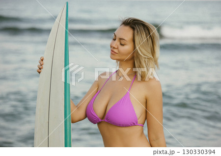 Attractive young woman in a purple bikini posing with a surfboard on the beach, enjoying summer by the sea, confident and relaxed, ocean waves in the background. 130330394