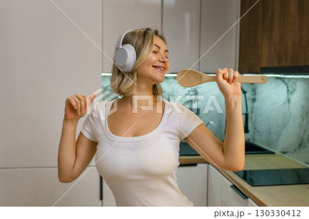 Happy young woman singing with wooden spoon like microphone, wearing headphones in modern kitchen. Joyful moment, music and fun at home. Happy young woman singing with wooden spoon like microphone, wearing headphones in modern kitchen. Joyful moment, music and fun at home. 130330412