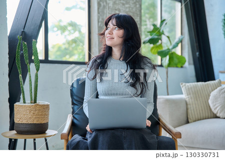 Portrait of young woman sitting on chair typing on laptop, in home interior Portrait of young woman sitting on chair typing on laptop, in home interior 130330731