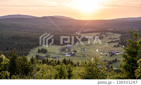 As the sun sets over Jizerka Village in the Jizera Mountains, the landscape glows with warm hues. The peaceful village rests amidst rolling hills and dense forests, evoking a sense of calm. 130331247