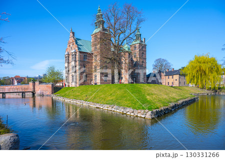 Rosenborg Castle showcases its stunning renaissance architecture in Copenhagen, surrounded by tranquil waters and lush greenery on a bright, clear day with blue skies. 130331266