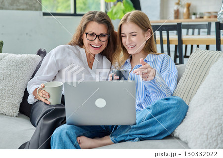 Middle aged mother and daughter 18,19 years old sitting together at home looking at laptop Middle aged mother and daughter 18,19 years old sitting together at home looking at laptop 130332022