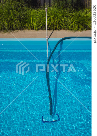 Pool vacuum cleaner with hose and telescopic pole submerged in clear blue water, shown during routine swimming pool maintenance on a sunny day. Pool vacuum cleaner with hose and telescopic pole submerged in clear blue water, shown during routine swimming pool maintenance on a sunny day. 130332620