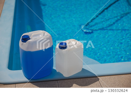 Two plastic chemical canisters with blue caps placed on the edge of a swimming pool, used for water treatment and maintenance in bright sunlight. 130332624