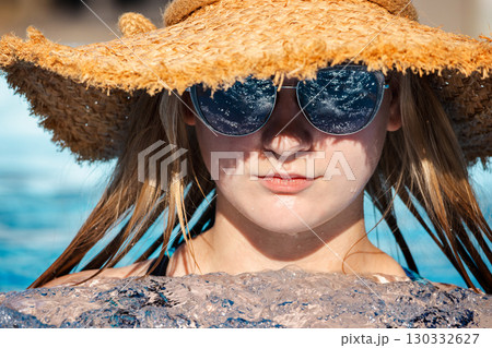 Smiling teenage girl cooling off in a bubbling pool, wearing reflective sunglasses and a wide straw hat under the bright summer sun 130332627