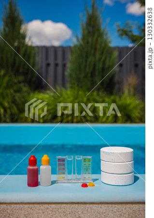 Chlorine tablets and a pool water test kit with chemical dropper bottles and color scale are arranged on the poolside in sunlight, used for pool maintenance. 130332638