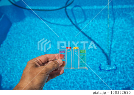 Hand holding a pool water test kit with pH and chlorine scale above a clear blue swimming pool, showing tools used for pool maintenance and water quality control. 130332650