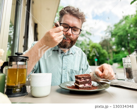 Man eating cake at outdoor cafe with tea pot and cup. Lifestyle and leisure dining experience of everyday relaxation and authentic food enjoyment. Man eating cake at outdoor cafe with tea pot and cup. Lifestyle and leisure dining experience of everyday relaxation and authentic food enjoyment. 130334348