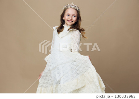 Happy girl in a white dress and crown, smiling on a neutral beige background, perfect for children s fashion and joyful moments 130335965