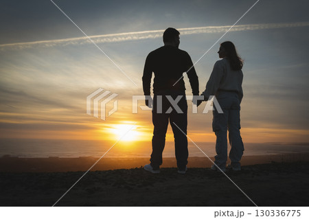 Couple holding hands watching golden sunset over ocean from hilltop 130336775
