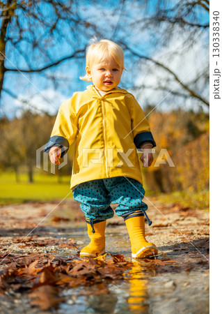 Sun always shines after the rain. Small bond infant boy wearing yellow rubber boots and yellow waterproof raincoat walking in puddles in city park on sunny rainy day 130338340