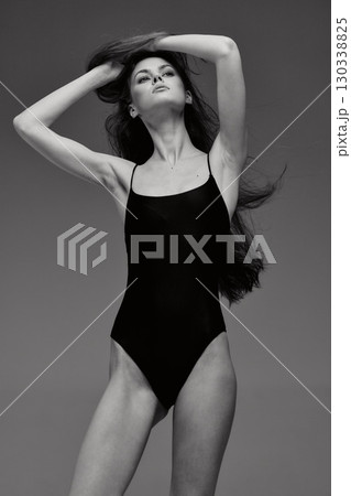 Black and white portrait of a tall slender woman posing confidently in a black swimsuit with flowing hair against a plain background. Black and white portrait of a tall slender woman posing confidently in a black swimsuit with flowing hair against a plain background. 130338825