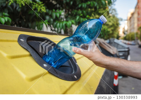 Hand recycling a blue plastic bottle into a yellow container on a city street in Rome, Italy, promoting waste sorting and environmental care. Hand recycling a blue plastic bottle into a yellow container on a city street in Rome, Italy, promoting waste sorting and environmental care. 130338993