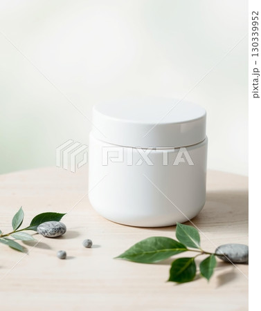 Minimalist white jar with lid, sitting on a light wooden surface with green leaves and small rocks. 130339952