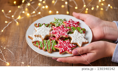 Christmas plate of festive cookies in snowflake and tree shapes with red, white and green icing held in hands on wooden background with lights 130340142