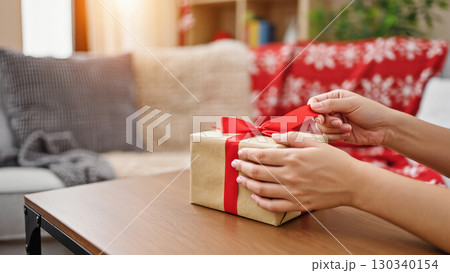 Woman's hands tying red ribbon on Christmas gift box on cozy home background with festive decor Woman's hands tying red ribbon on Christmas gift box on cozy home background with festive decor 130340154