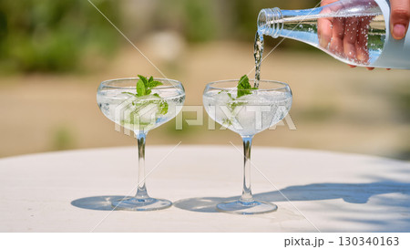 Two refreshing summer cocktails with ice and mint in coupe glasses, with sparkling water being poured on sunny outdoor table. Two refreshing summer cocktails with ice and mint in coupe glasses, with sparkling water being poured on sunny outdoor table. 130340163