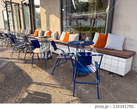 Outdoor cafe terrace with blue chairs, small tables, and pillows in sunlight. Hospitality, lifestyle, and social culture expressed through urban leisure and modern dining. 130341780