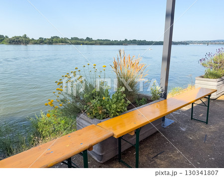 Wooden bench near riverbank with flowers and calm water under blue sky. Leisure, recreation, and urban nature experience through riverside landscape and outdoor lifestyle. Wooden bench near riverbank with flowers and calm water under blue sky. Leisure, recreation, and urban nature experience through riverside landscape and outdoor lifestyle. 130341807