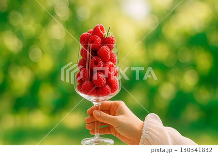 A hand holds a wine glass filled with fresh raspberries against a blurred green outdoor background. Generated AI. 130341832