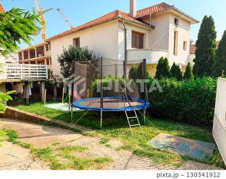 Backyard with trampoline near house. Family lifestyle, play, and suburban residential environment with construction site view. Backyard with trampoline near house. Family lifestyle, play, and suburban residential environment with construction site view. 130341912