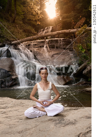 Young woman sitting in lotus pose under tropical waterfall meditating with calm strength and mindfulness fully connected to nature energy tranquility and inner balance Young woman sitting in lotus pose under tropical waterfall meditating with calm strength and mindfulness fully connected to nature energy tranquility and inner balance 130342611