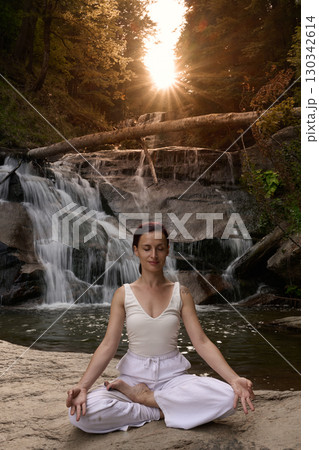 Young woman sitting in lotus pose under tropical waterfall meditating with calm strength and mindfulness fully connected to nature energy tranquility and inner balance Young woman sitting in lotus pose under tropical waterfall meditating with calm strength and mindfulness fully connected to nature energy tranquility and inner balance 130342614