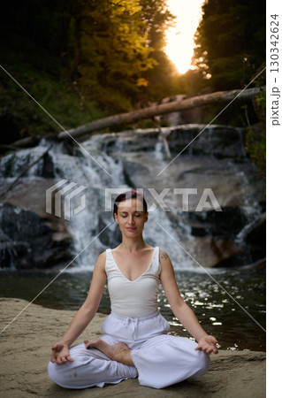 Young woman sitting in lotus pose under tropical waterfall meditating with calm strength and mindfulness fully connected to nature energy tranquility and inner balance Young woman sitting in lotus pose under tropical waterfall meditating with calm strength and mindfulness fully connected to nature energy tranquility and inner balance 130342624
