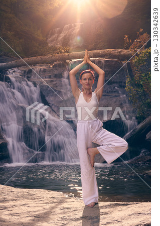 Woman doing yoga tree pose in front of waterfall at sunrise peaceful meditation in nature Woman doing yoga tree pose in front of waterfall at sunrise peaceful meditation in nature 130342639