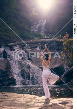 Woman doing yoga tree pose in front of waterfall at sunrise peaceful meditation in nature  130342640