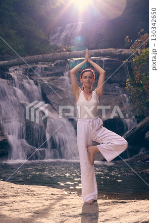Woman doing yoga tree pose in front of waterfall at sunrise peaceful meditation in nature Woman doing yoga tree pose in front of waterfall at sunrise peaceful meditation in nature 130342650