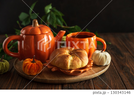 Autumnal tea set with pumpkin-shaped teapot and mug, and a baked bun on a wooden table. Autumnal tea set with pumpkin-shaped teapot and mug, and a baked bun on a wooden table. 130343146