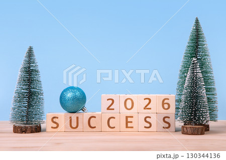 New Year 2026 and Christmas postcard. Wooden cubes with numbers 2026 and word success and green Christmas trees on a wooden table and a blue background. 130344136