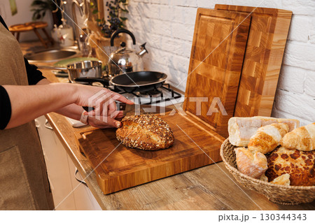 Woman slicing multigrain bread on wooden cutting board in cozy home kitchen Woman slicing multigrain bread on wooden cutting board in cozy home kitchen 130344343