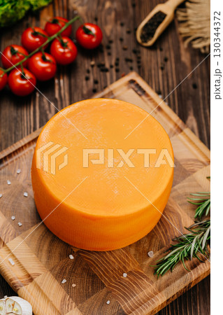 Whole round cheese wheel on wooden board with herbs and vegetables on orange background Whole round cheese wheel on wooden board with herbs and vegetables on orange background 130344372
